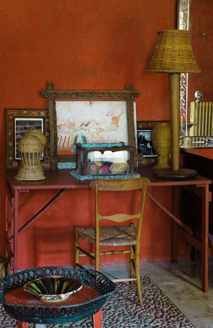 Wooden chair and vintage desk decorated with pictures and wicker ornaments against wall painted rusty red