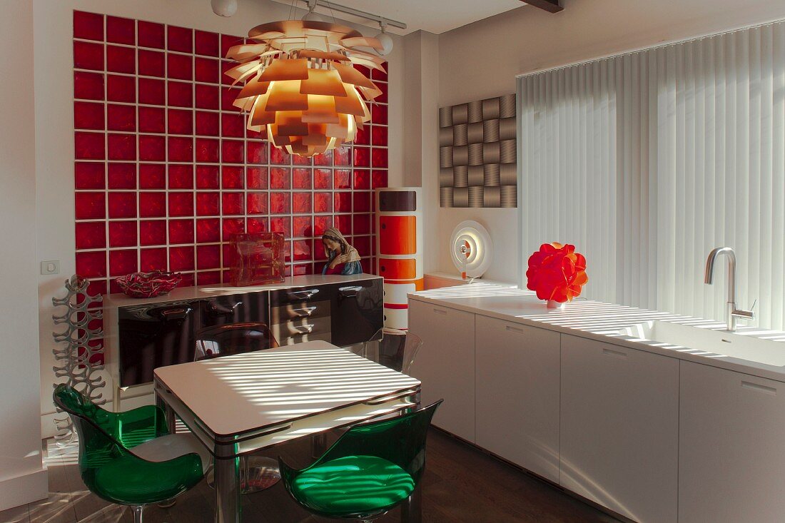 White kitchen counter below vertical louvre blinds, dining table and chairs below classic pendant lamp and retro sideboard against wall covered in red glass tiles