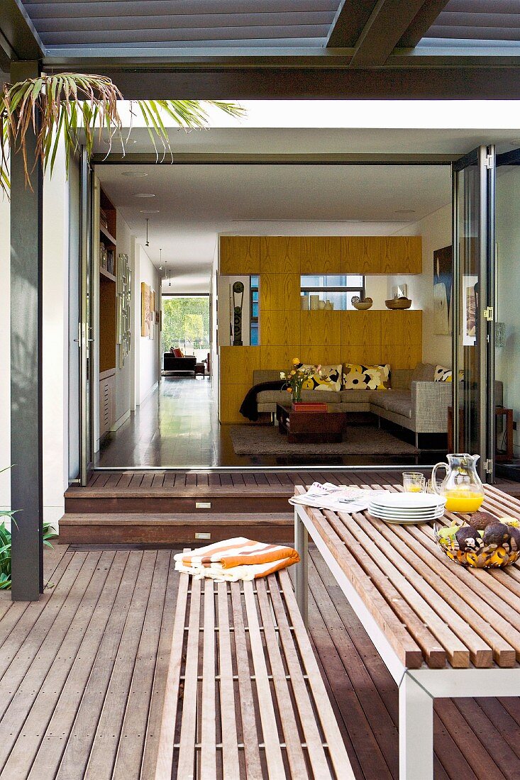 Table and bench on wooden terrace in front of open sliding folding doors with view into living room