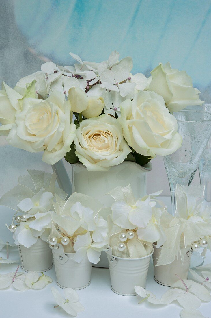Weisser Blumenstrauss aus Hortensienblüten und Rosen im Porzellankrug, davor Hochzeitsmandeln mit weissen Schleifen in kleinen Eimern