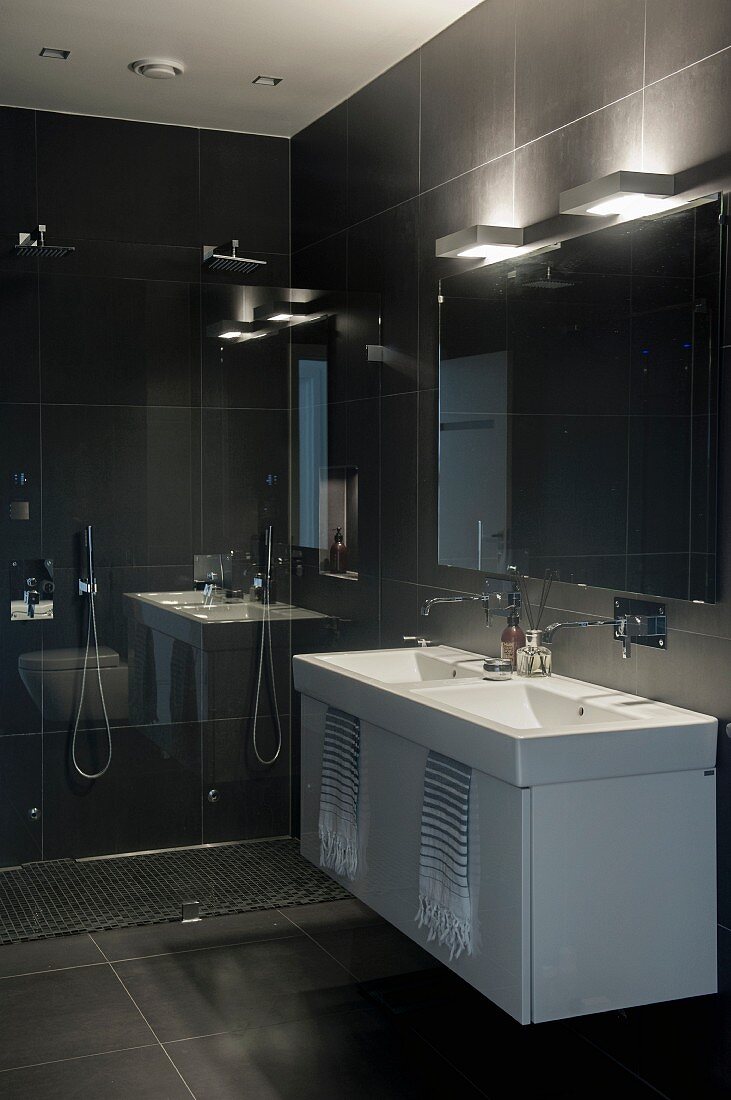 Washstand with white base cabinet against black-tiled wall below wall lamps above mirror in designer bathroom