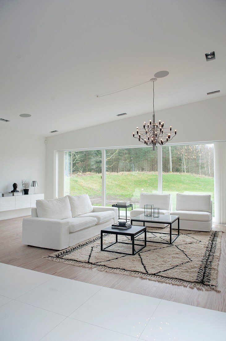 Elegant white two-seater sofa and matching armchair around two coffee tables on rug below chandelier; view of garden through large window