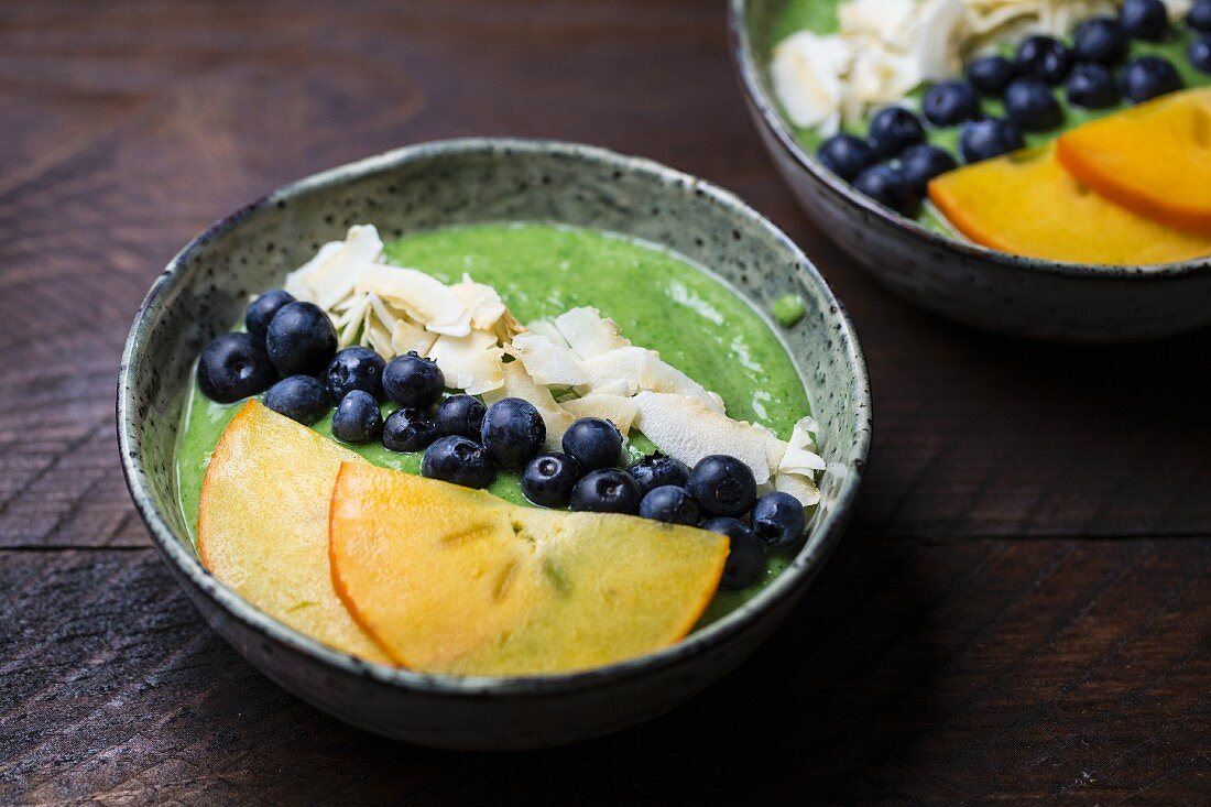 Grüner Smoothie im Schälchen mit Blaubeeren, Kokosflocken und Kakischeiben