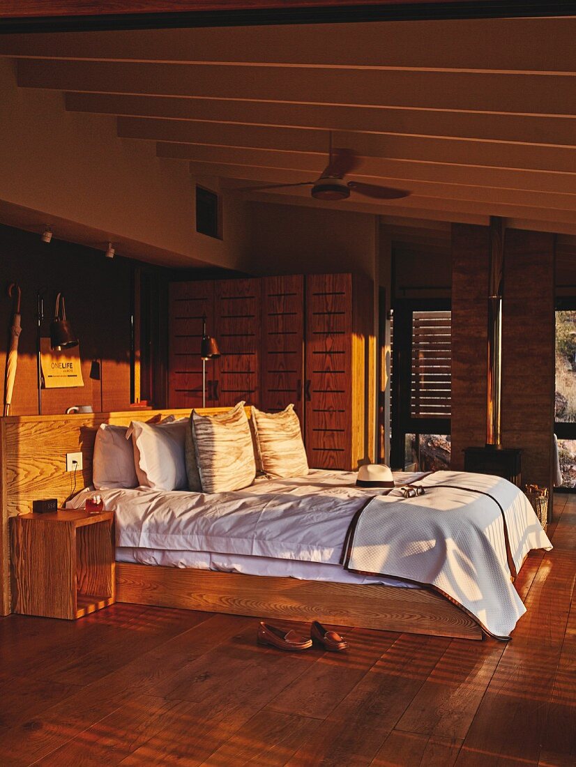 Double bed with wooden frame and sun shining on headboard in bedroom in shades of brown