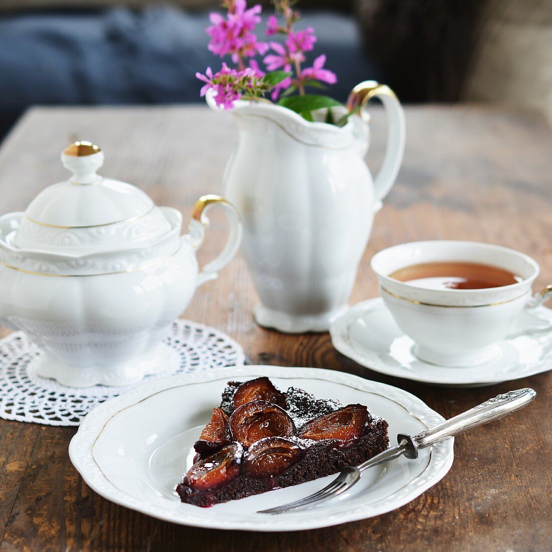 A slice of chocolate cake with plums and icing sugar served with tea