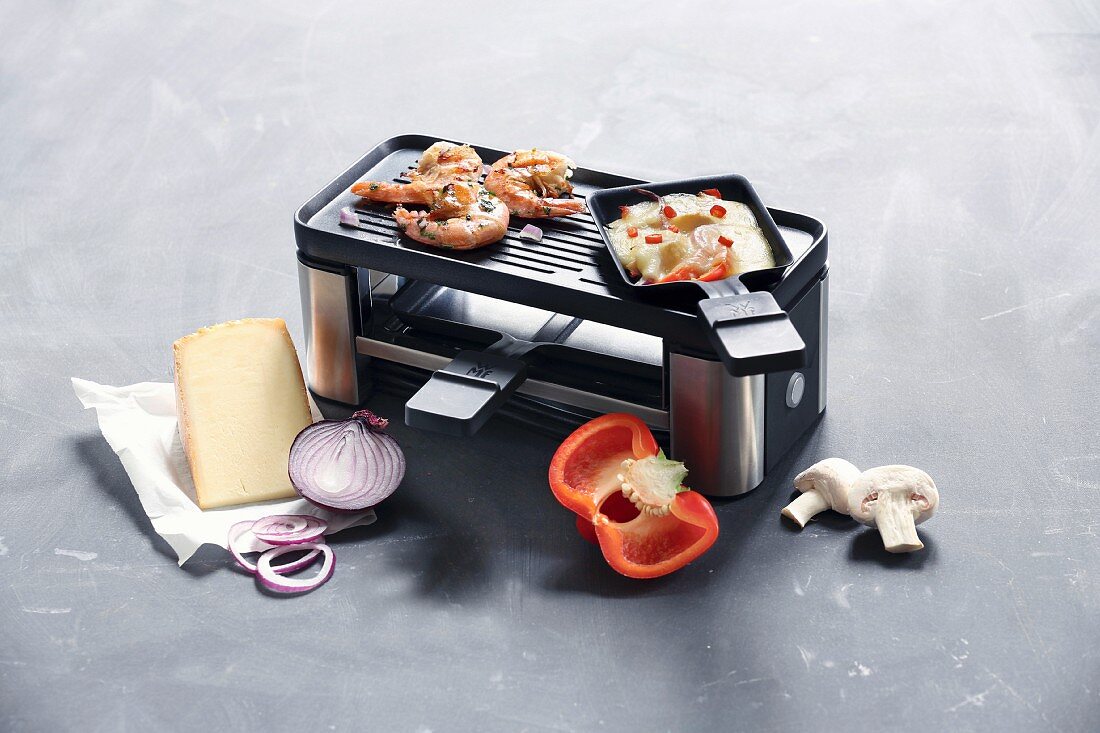 A raclette for two pans with a grill surface surrounded by ingredients for raclette