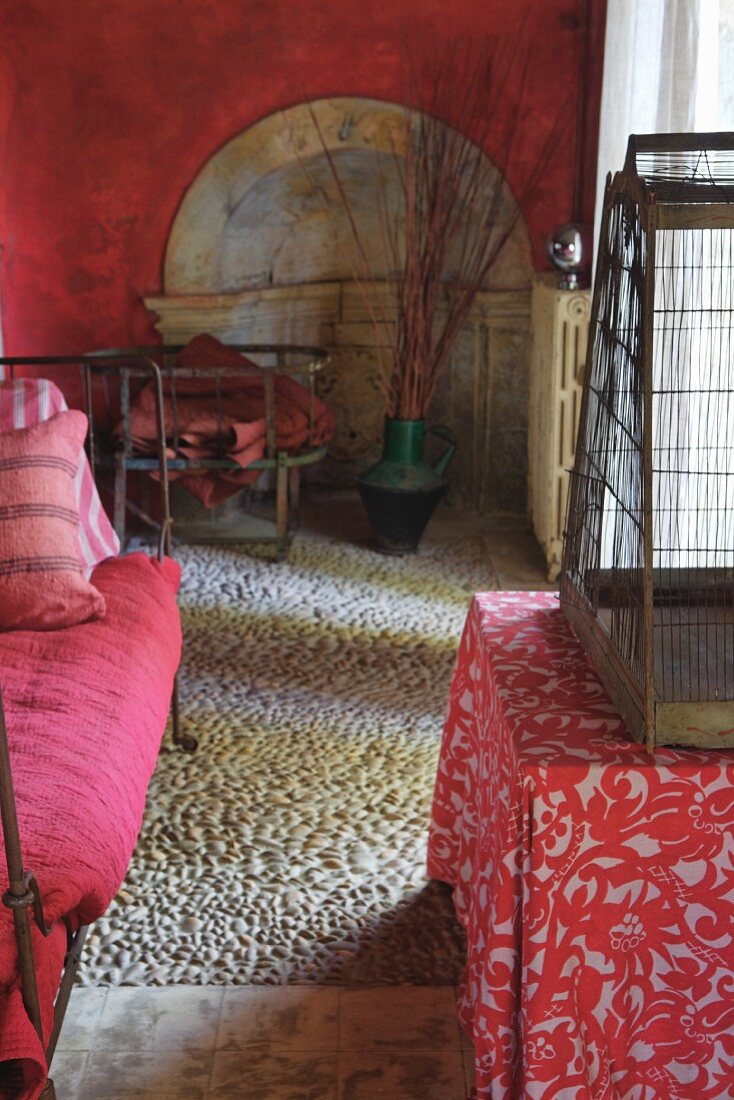 Mediterraner Wohnraum in Rot mit Steckkieselboden, Bett und Tisch mit floral gemusterter Tischdecke und Vogelkäfig