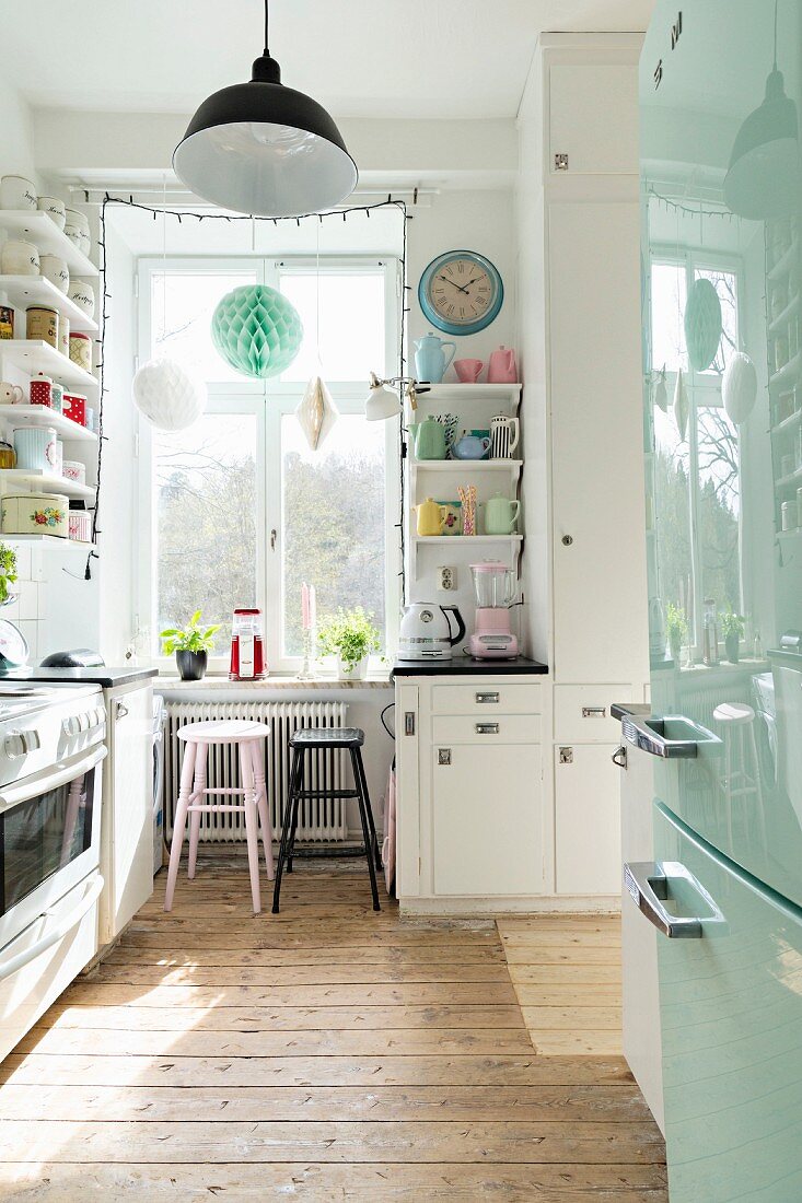 Kitchen with rustic wooden floor, pale blue fridge, bar stools at counter below window and open-fronted shelving