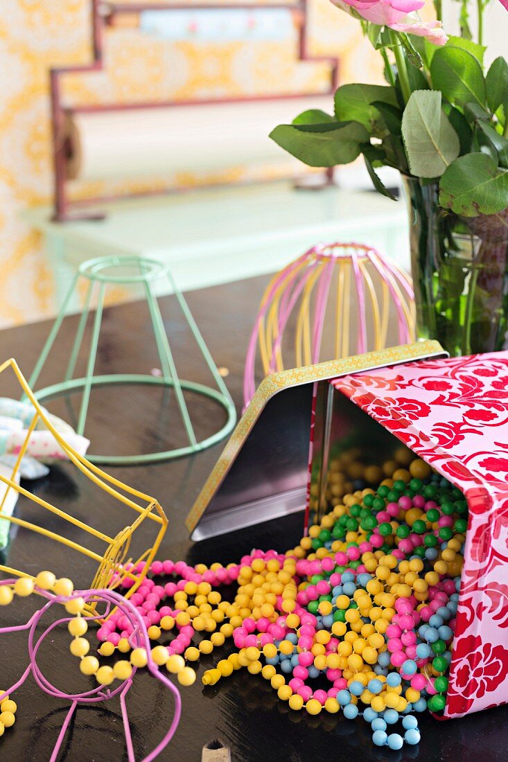 Strings of colourful plastic beads spilling out of open tin and lampshade frames made from pastel wire on tabletop