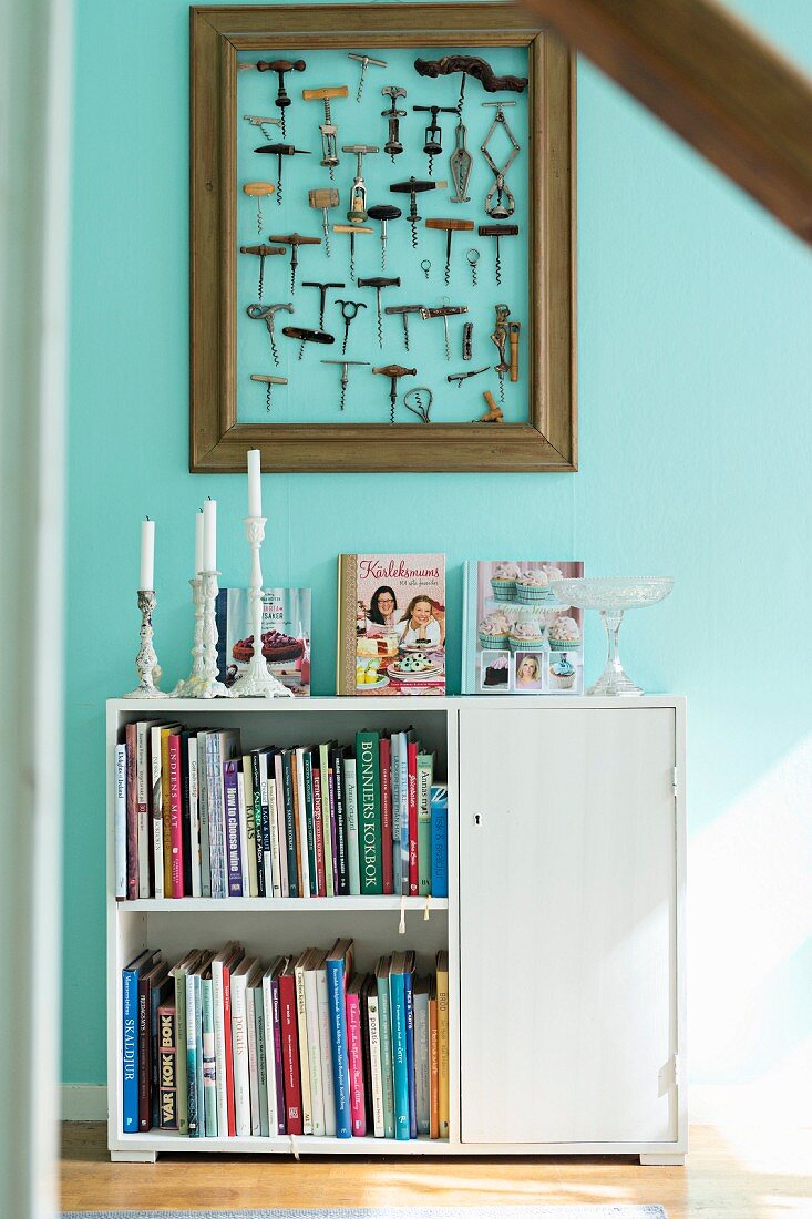 White half-height cabinet with books on shelves and candles on top against turquoise wall below framed collection of corkscrews