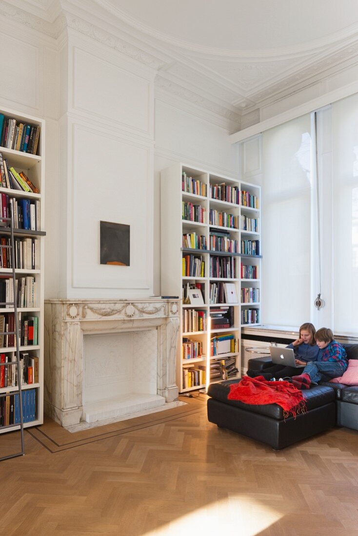 Stucco ceiling, children using laptop sitting on black sofa and bookcases in living room in period apartment