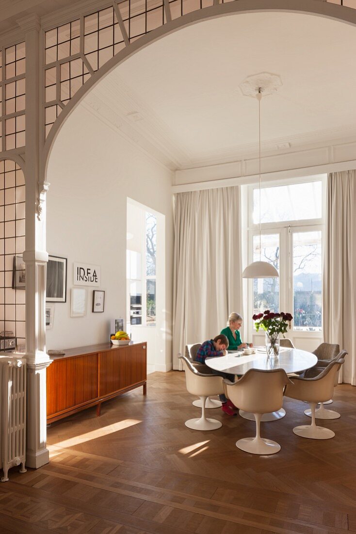 Blick durch Rundbogen-Durchgang mit Bleiverglasung auf eleganten Essplatz mit Designklassikern, Kind und Mutter am Tisch