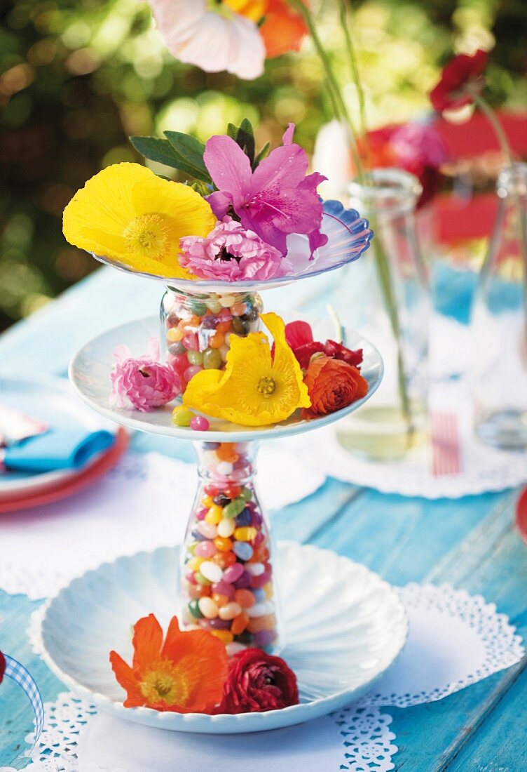 A tiered stand made of plates and bottles filled with colourful sweets, decorated with fresh flowers on a summery set table