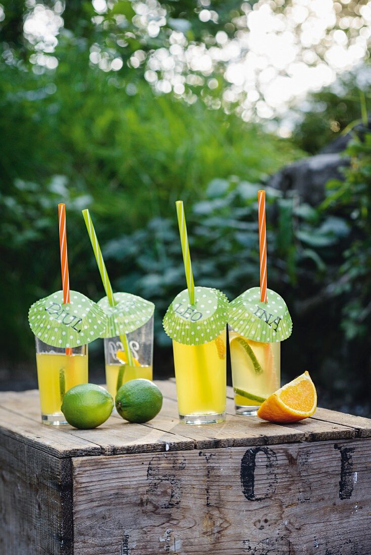 Refreshing drinks decorated with straws and names written on paper covers arranged with limes and orange wedge on wooden crate
