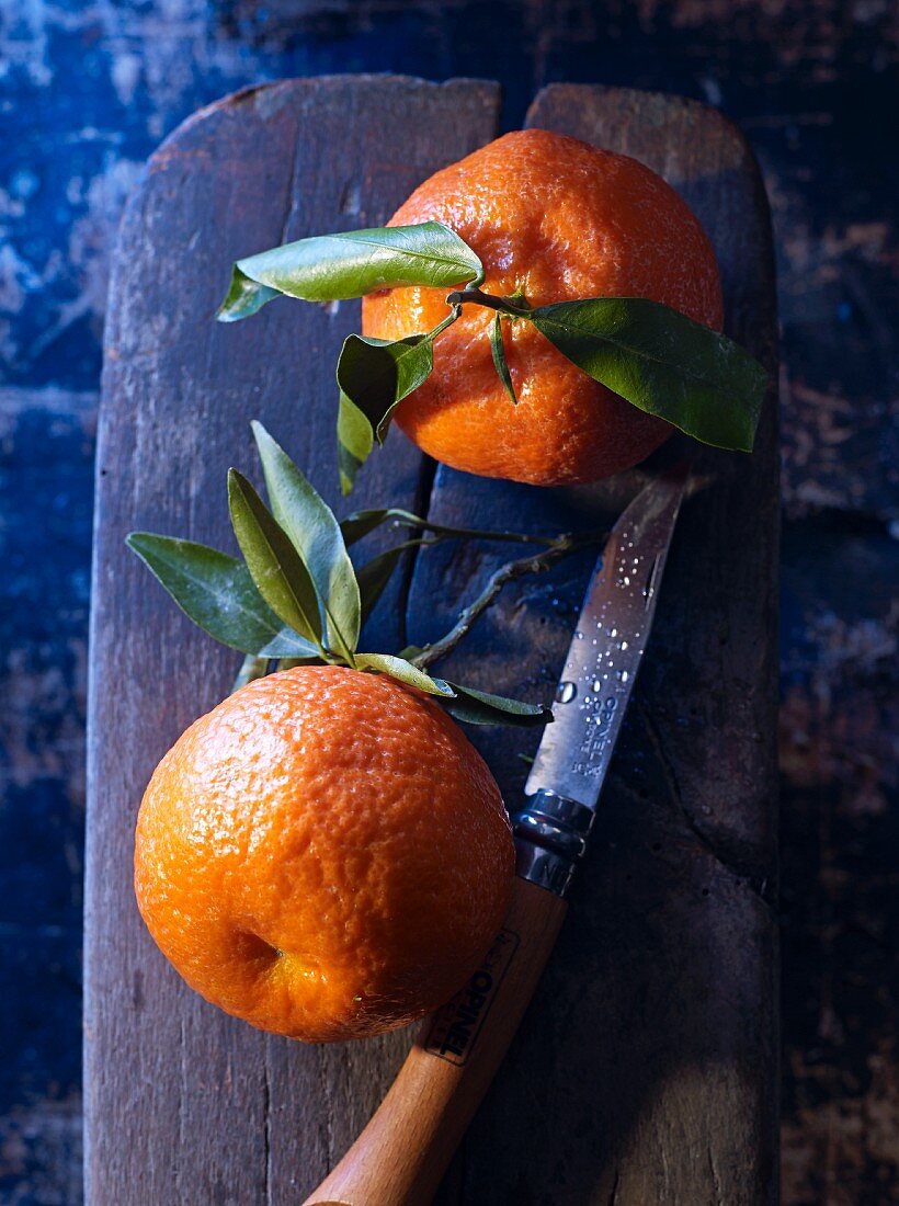 Zwei Clementinen mit Blättern auf Schneidebrett mit Messer