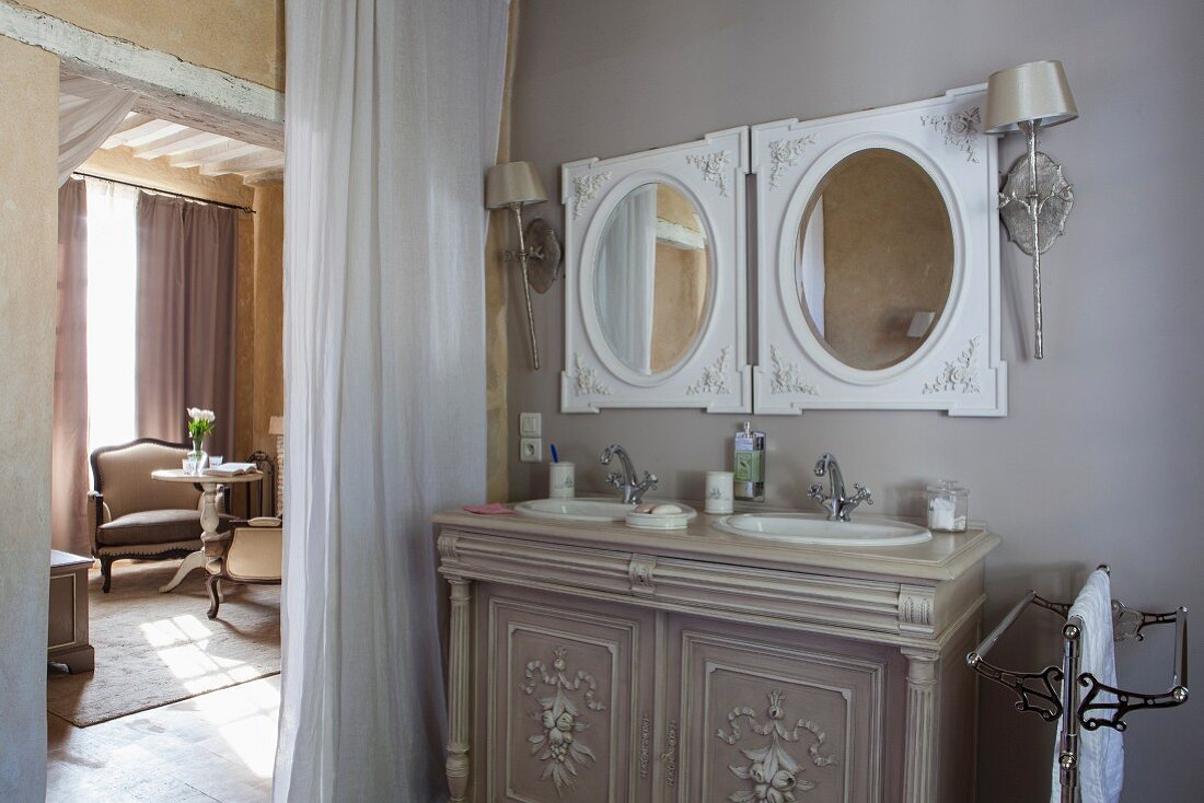 Elegant washstand with twin inset sinks below round mirrors next to curtain in doorway