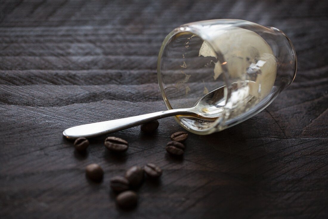 Leergetrunkenes Espressoglas mit Löffel & Kaffeebohnen auf Holzuntergrund