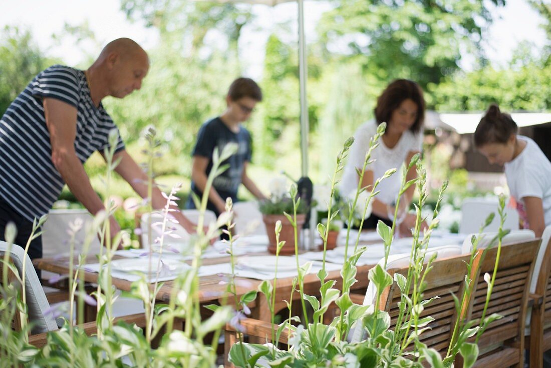 Blick über Grünpflanze auf Familie beim Brunch vorbereiten in sommerlichem Garten