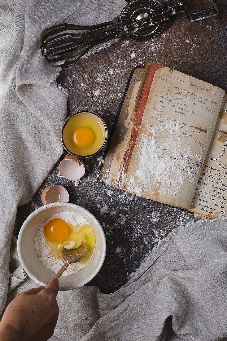 A baking scene with eggs, flour, a recipe book and a whisk