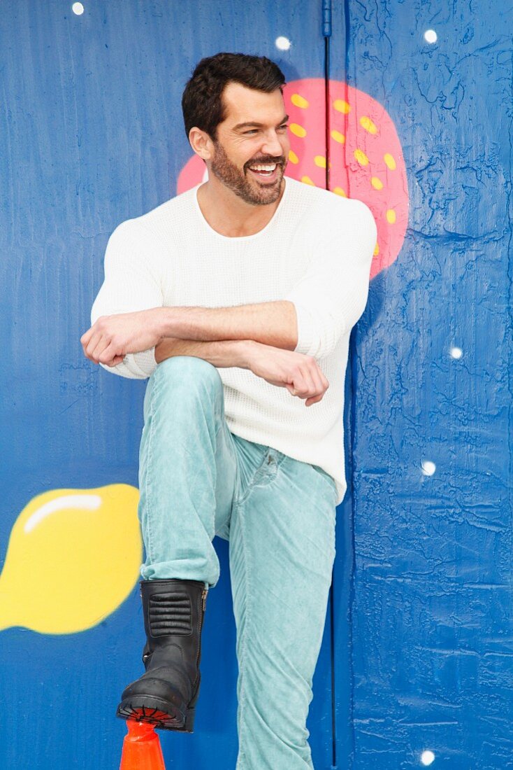 A man wearing a white jumper and jeans leaning against a painted wall