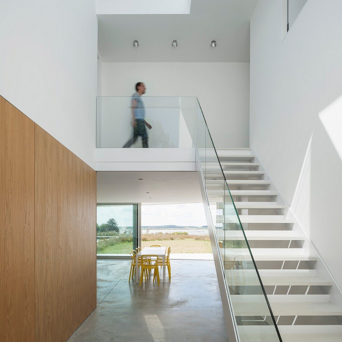 Treppe mit Glasgeländer in minimalistischem Wohnbereich, im Hintergrund Esstisch vor Terrassenschiebetüren