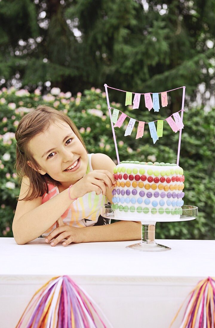A girl in a garden next to a birthday cake decorated with colourful chocolate beans