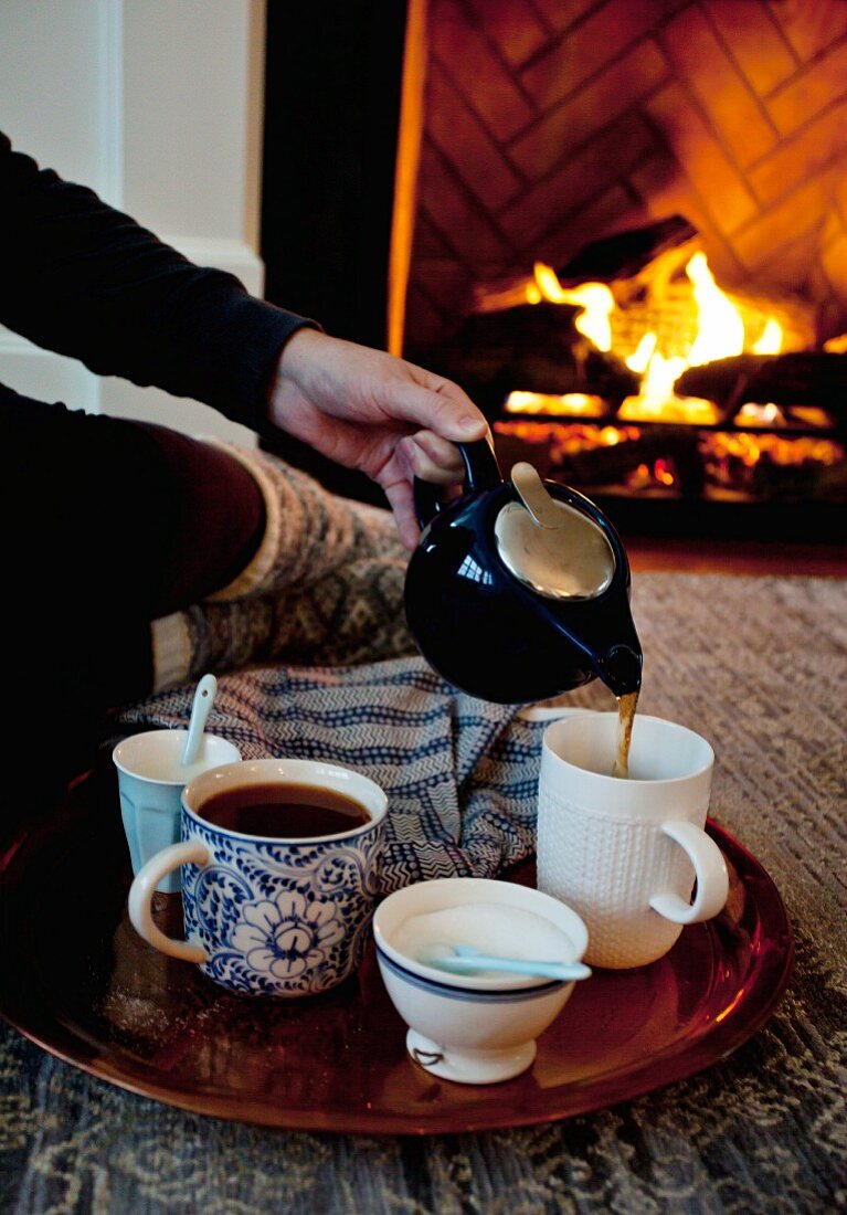 A woman sitting by a fireplace pouring tea into a cup