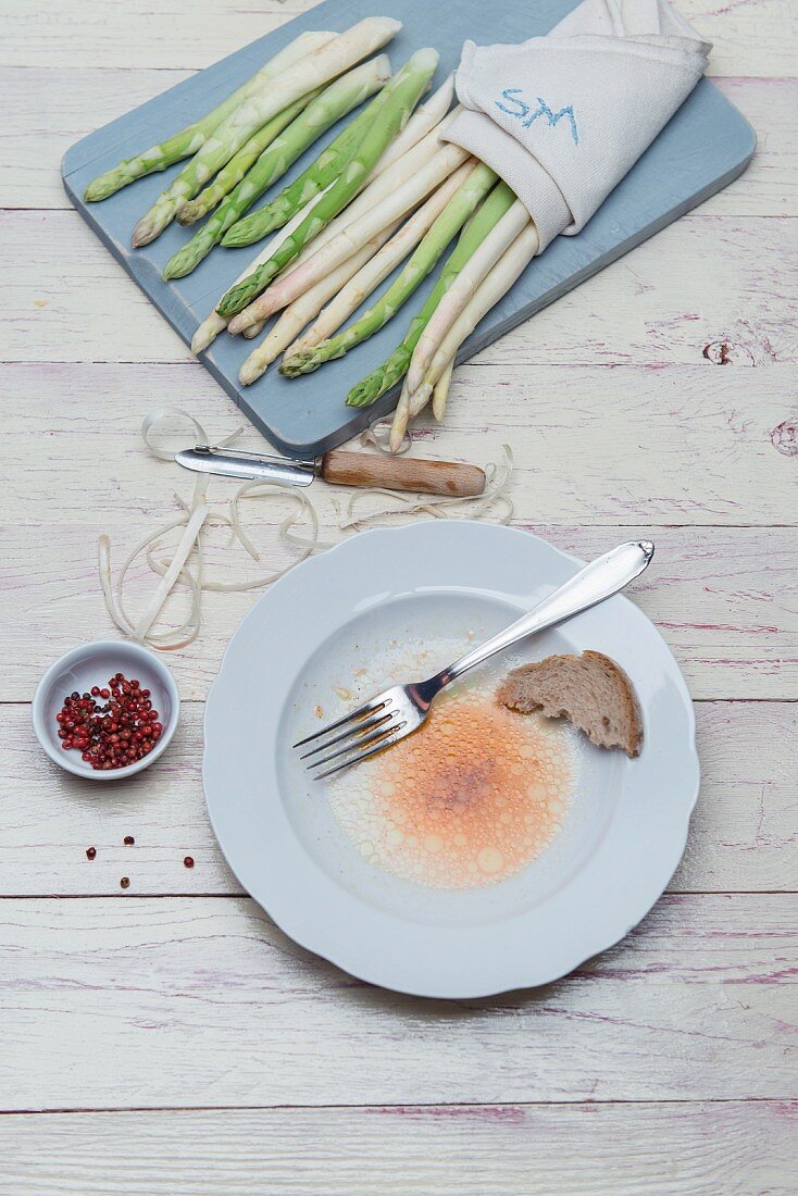 White and green asparagus on a wooden board with peelings and a peeler, the remains of vinaigrette on a plate with a piece of bread and a fork