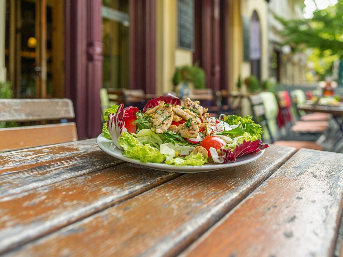 A mixed leaf salad with tomatoes and radishes on a wooden table outside a restaurant