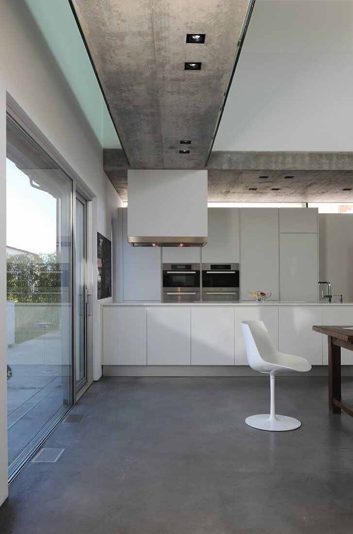 Open-plan kitchen, white classic shell chair and recessed spotlights in exposed concrete ceiling in modern house