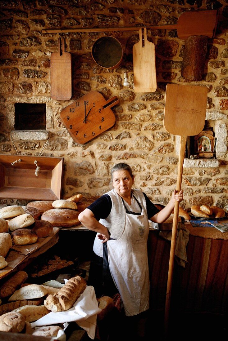 Baker with bread shovel next to counter with fresh loaves of bread in bakery with rustic natural stone wall