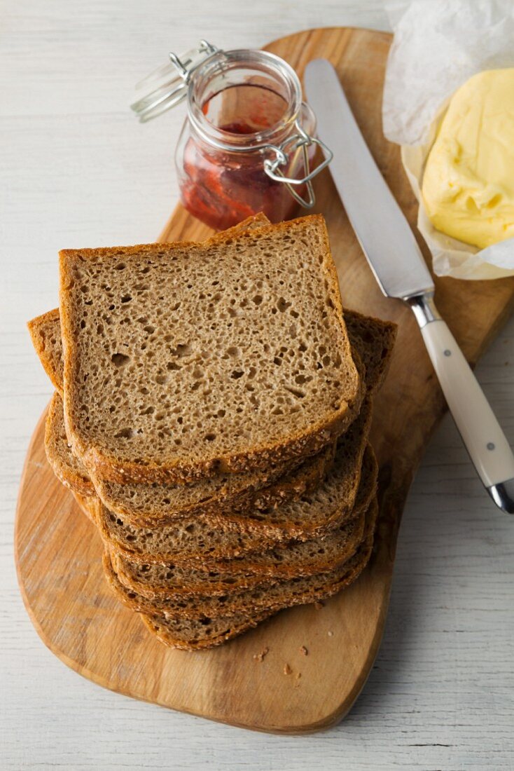 Slices of wholemeal bread stacked on a wooden board with butter and strawberry jam