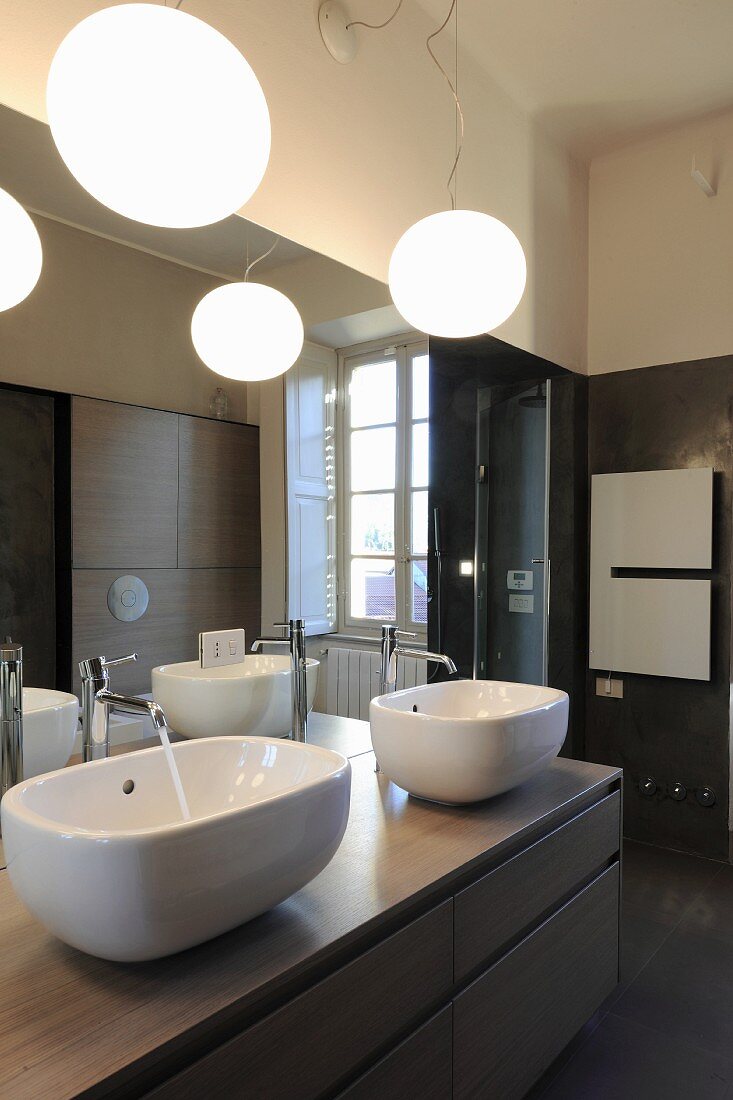 Zwei weiße Waschschüsseln auf Waschtischmöbel vor grossflächigem Spiegel, oberhalb Kugelleuchten im Bad