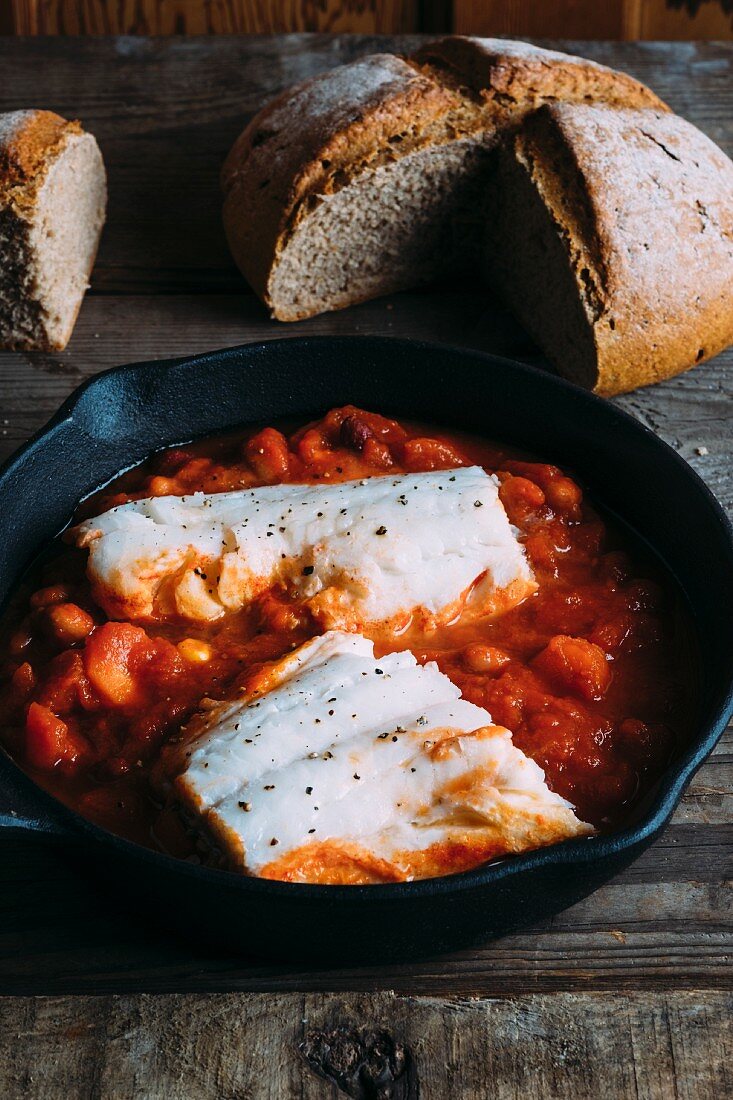 Tomato and bean stew with cod in a cast-iron pan with a loaf of rye bread in the background
