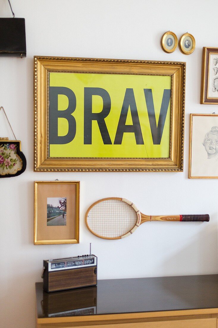 Gallery of gilt-framed pictures and tennis racquet hung on wall above retro portable radio on sideboard