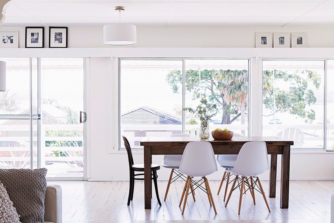 Open living area with wooden table and white shell chairs in front of the window front and picture gallery below the ceiling