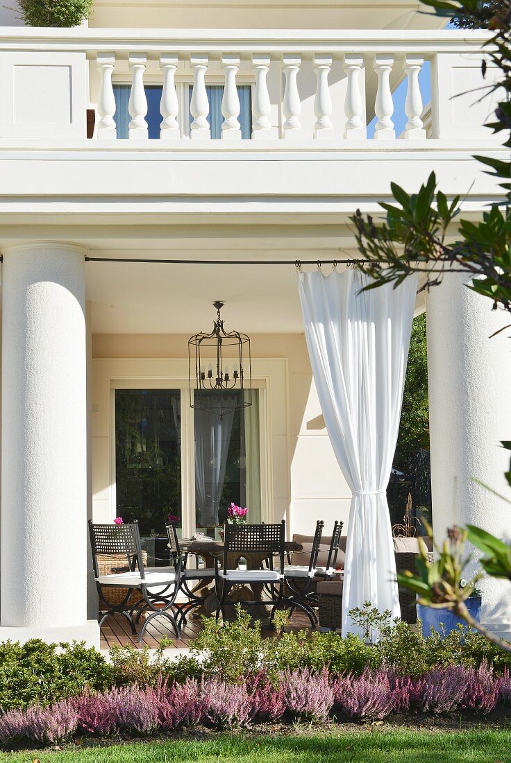 Veranda of villa with enormous pillars and gathered curtains below balcony with postmodern stone balustrade