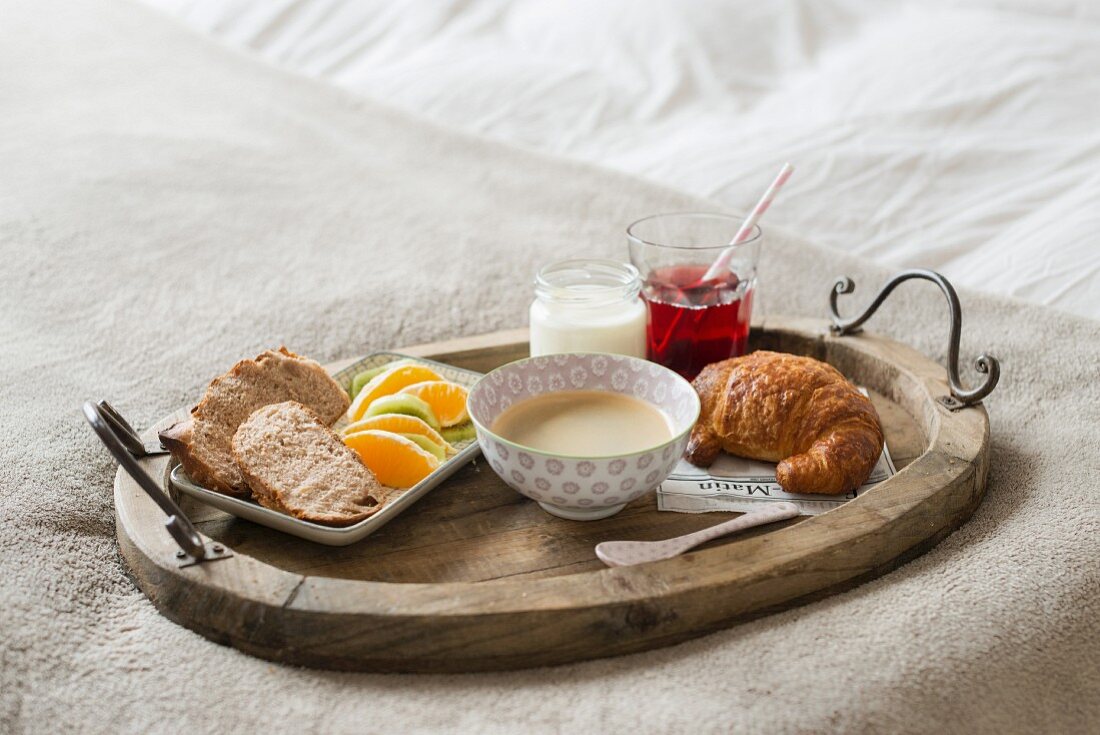 Frühstückstablett mit Milchkaffee, Croissant und frischem Obst auf weißem Bett
