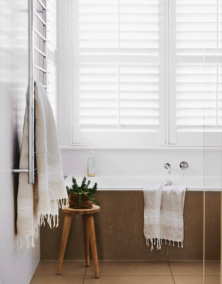 Stone-clad bathtub in reduced bathroom with white blinds and rustic wooden stool