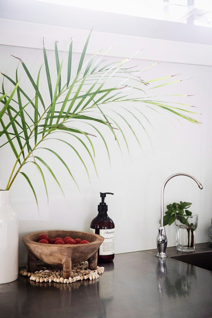 Kitchen worktop with gray polished surface, palm branches in vase and rustic wooden bowl next to soap dispenser at the sink