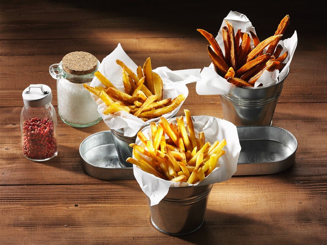 Drei verschiedene Sorten Pommes Frites mit Salzfass und rotem Pfeffer auf rustikaler Holzplatte