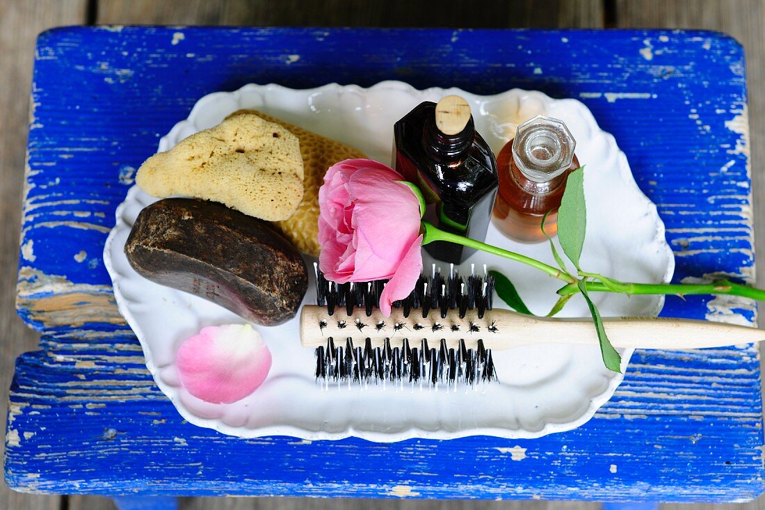 Weisses Tablett mit Schwamm, Badezubehör, Haarbürste und einer Rose auf blauem Holzbrett