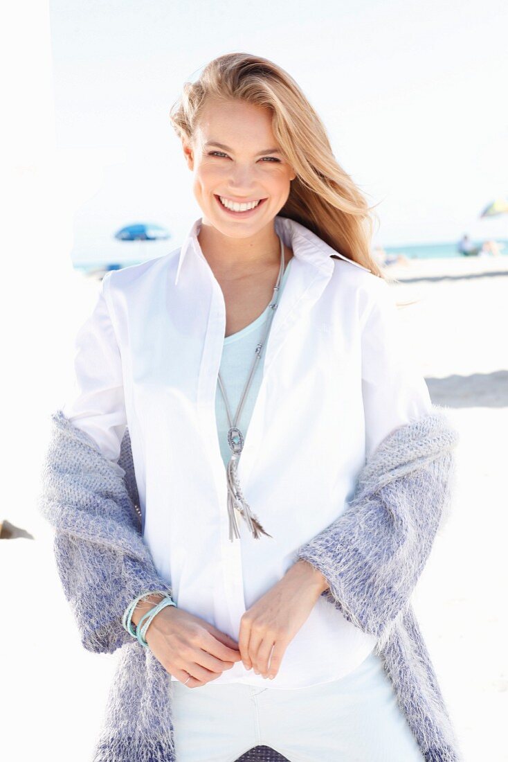 A blonde woman on a beach wearing a white blouse and a purple cardigan