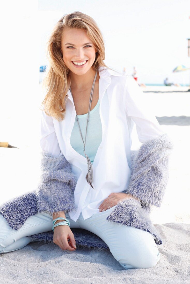 A blonde woman on a beach wearing a white blouse and a purple cardigan