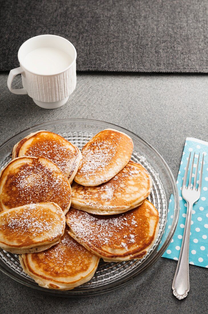 Buttermilch-Pancakes mit Puderzucker auf Glasteller vor einer Tasse Milch