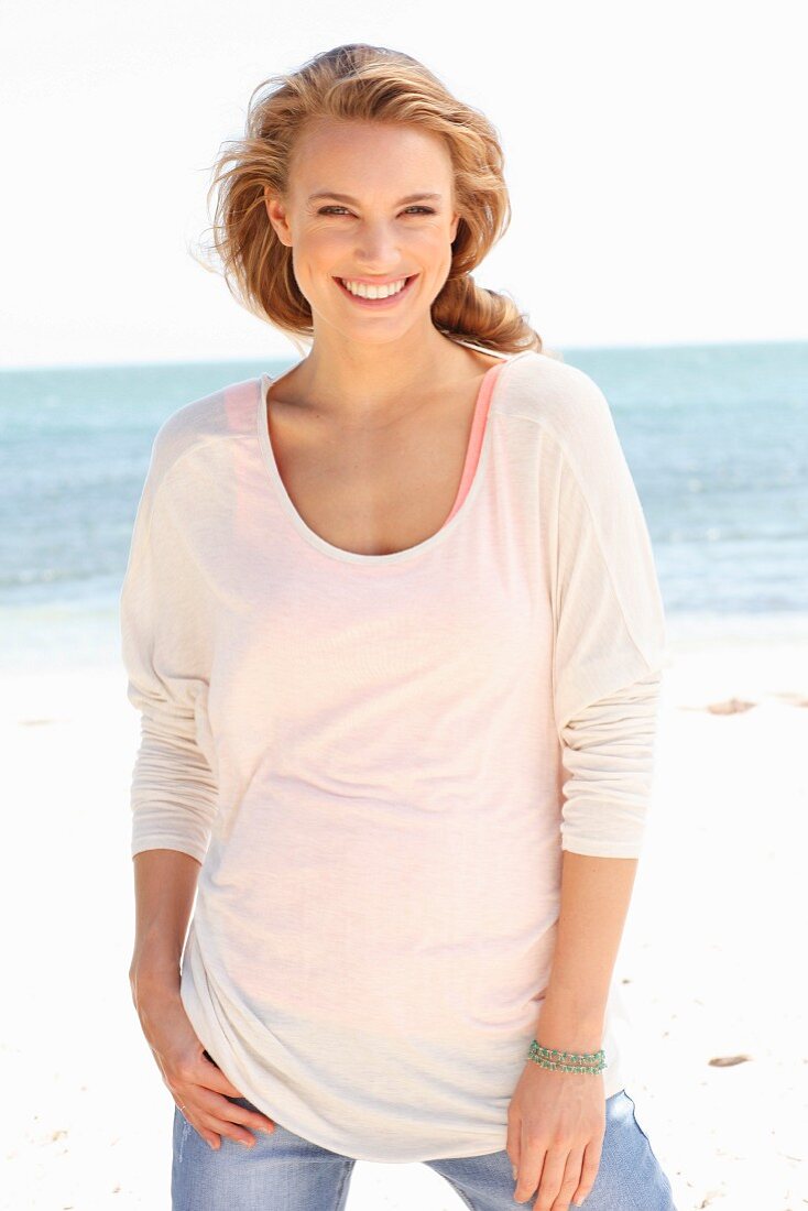 A blonde woman on a beach wearing a top, a thin, long-sleeved top and jeans