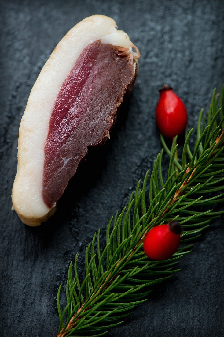 A slice of smoked, marinated duck breast with a sprig of spruce and rose hips on a slate platter