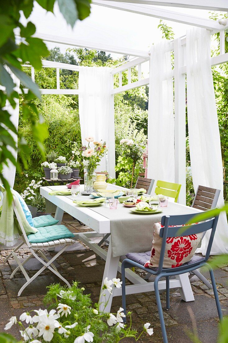 A table laid on a terrace with chairs with coloured cushions under a pergola with light curtains on a white frame