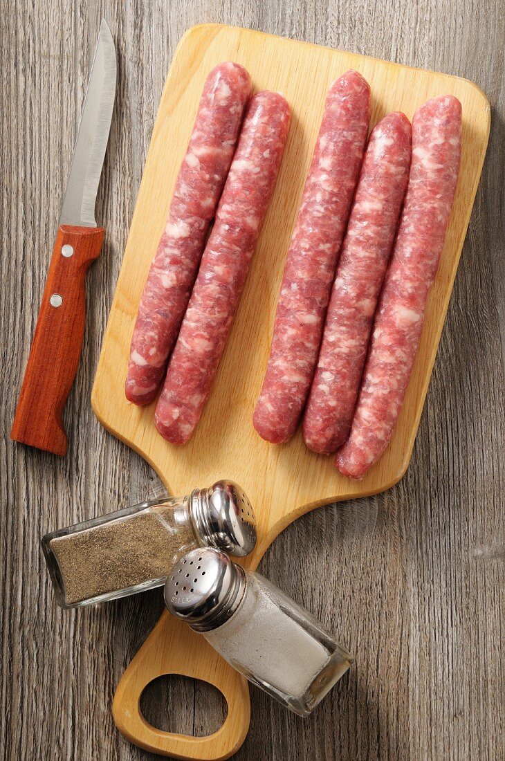 Chipolata-Würstchen auf Holzbrett mit Messer, Pfeffer und Salz