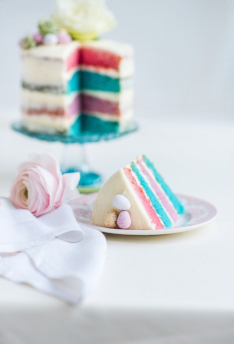 Vierschichtiger Rainbow Cake mit weißem Zuckerguss, Ostereiern und Blumendeko (angeschnitten)